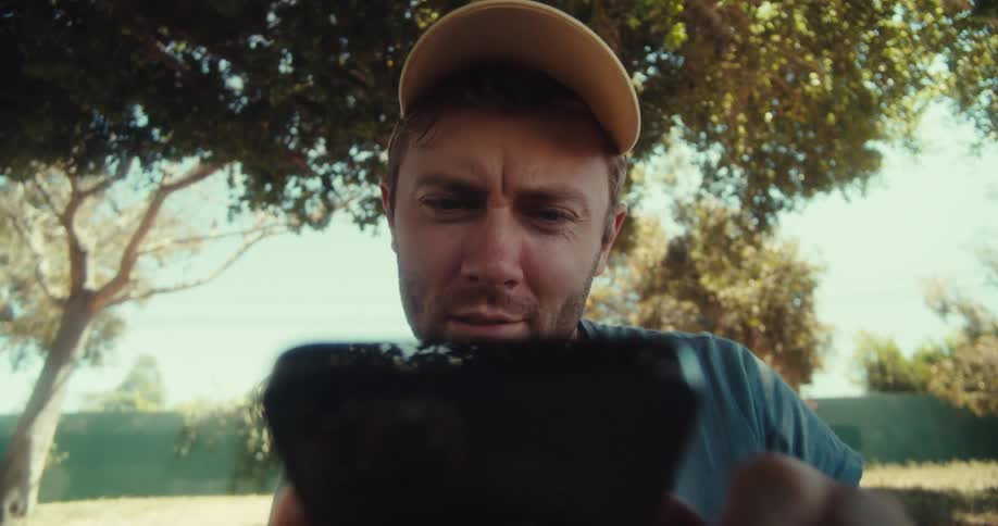 A man in a yellow cap and blue shirt sits outdoors under trees looking down while using his phone