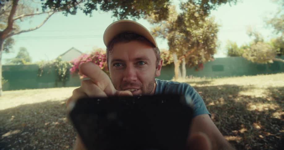 A man in a yellow cap and blue shirt sits in a park on a sunny day pointing with his finger and waving while holding a phone