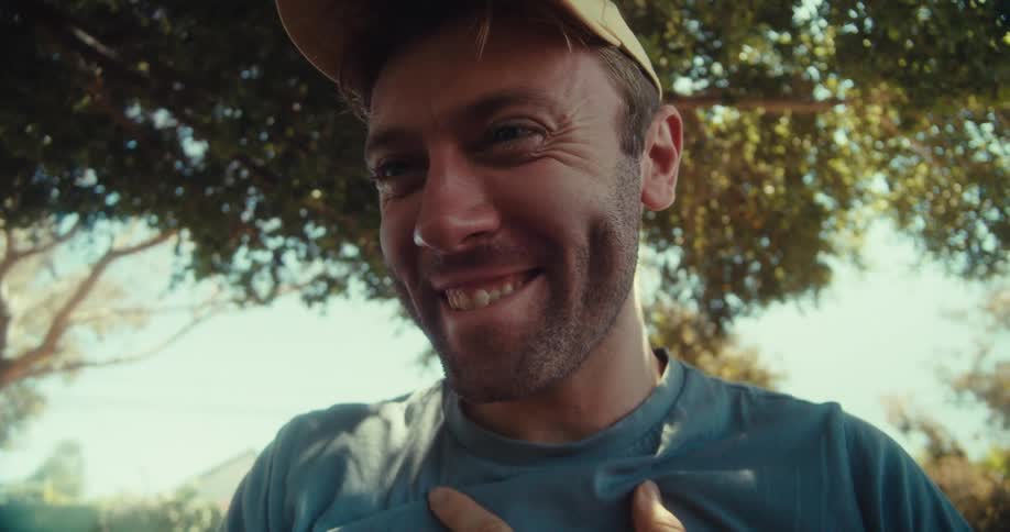 A man in a yellow cap and blue shirt sits in a park on a sunny day browsing his phone with a surprised expression