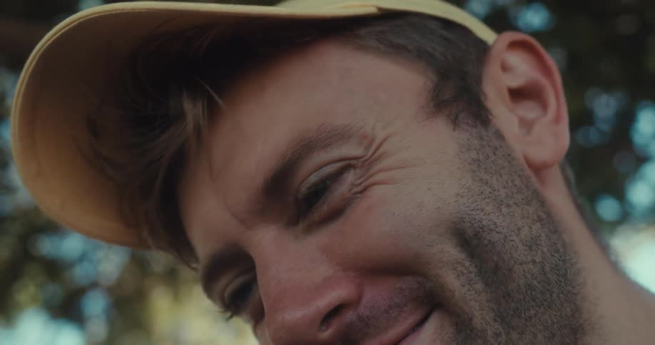 A man outdoors wearing a hat looks at his phone with a smile on his face at midday