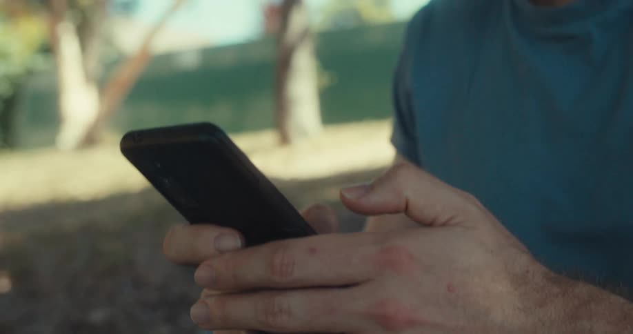 A man sits on the floor in a public park looking at his phone and smiling