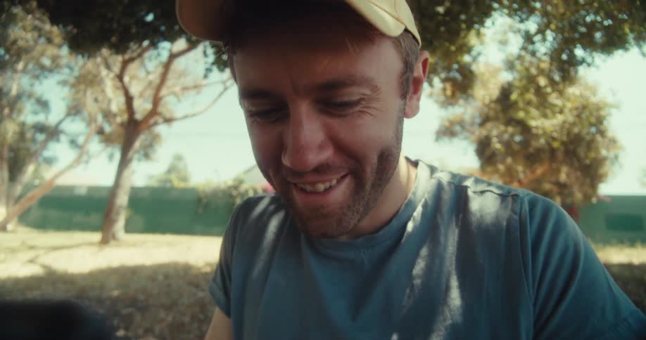 A man sits on the ground outdoors in a public park wearing a hat and smiling while looking at his phone