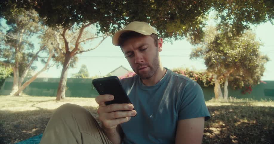 A man sits on the floor in a public park at midday scrolling through his phone smiling