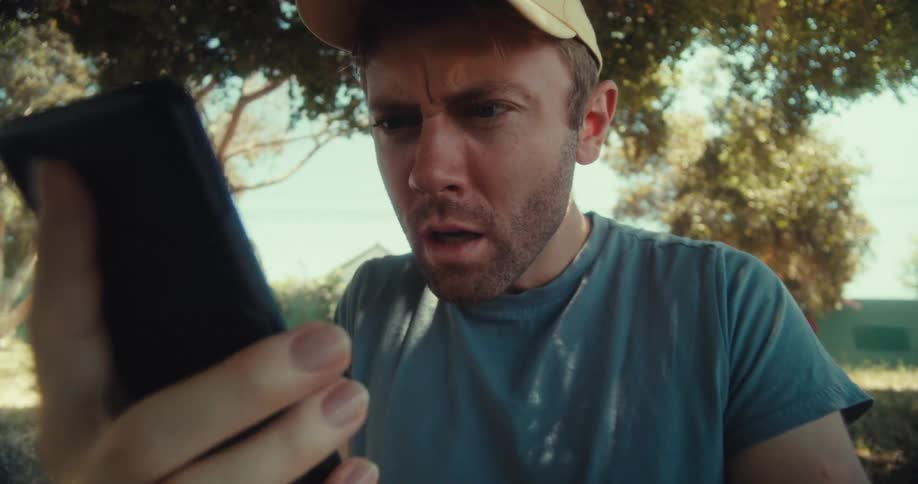 A man sits on the floor in a public park looking at his phone and reacting in disbelief