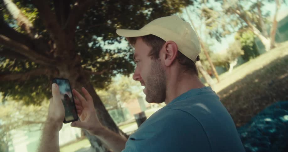 A man in a yellow cap and blue shirt lies on his back in a park sits down browsing his phone with a surprised expression on a sunny day