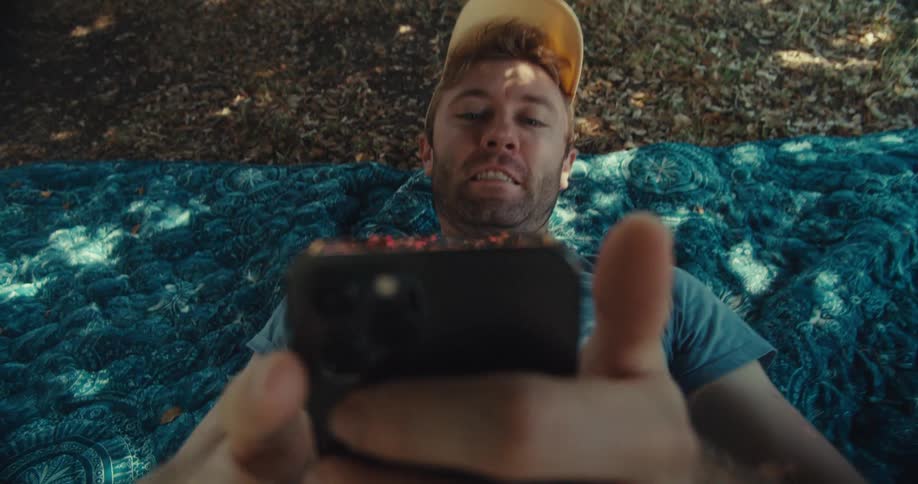 Man lies back on a blanket in a residential park while viewing his smartphone screen