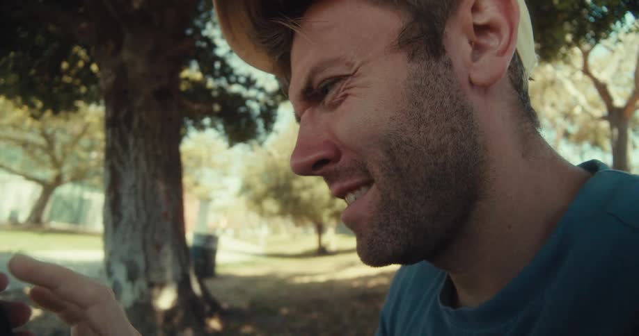 A man in a yellow cap and blue shirt lies on his back in a park browsing his phone with a confused expression on a sunny day