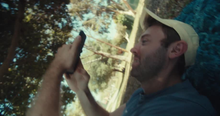 A man in a yellow cap and blue shirt sits in a park on a sunny day then lies down while browsing his phone with a happy expression