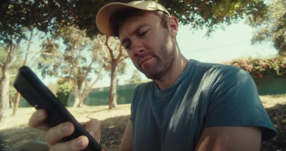 A man in a yellow cap and blue shirt sits outdoors under trees looking down while using his phone