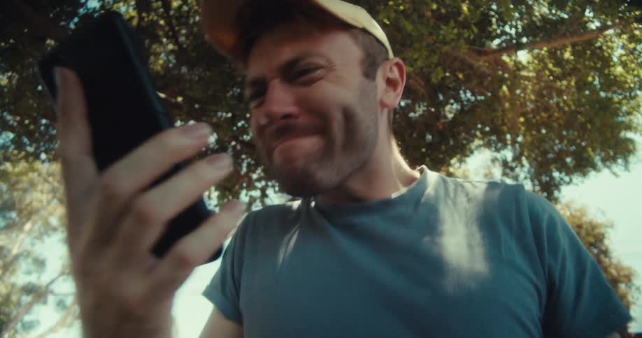 A man in a yellow cap and blue shirt sits in a park on a sunny day browsing his phone with an angered expression