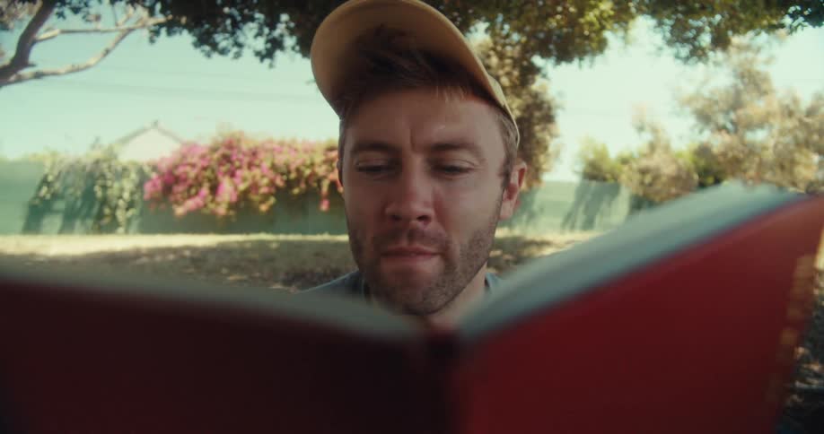 A man in a yellow cap and blue shirt sits in a park on a sunny day reading a book under a tree