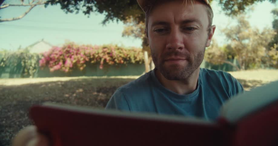 A man in a yellow cap and blue shirt sits in a park on a sunny day writing on a red notebook under a tree