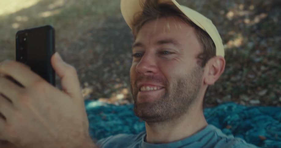 A man laughs while sitting in a park scrolling through his phone then lies down wearing a hat at midday