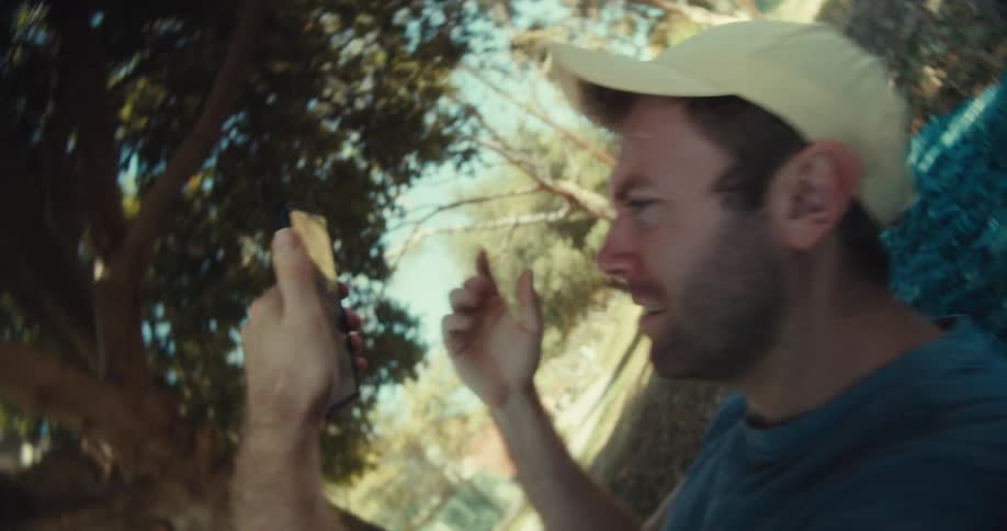 A man in a yellow cap and blue shirt sits in a park on a sunny day then lies down while browsing his phone with a confused expression
