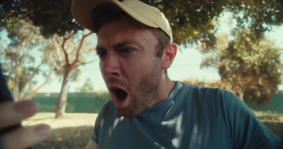 A man in a yellow cap and blue shirt sits in a park on a sunny day browsing his phone with a surprised expression