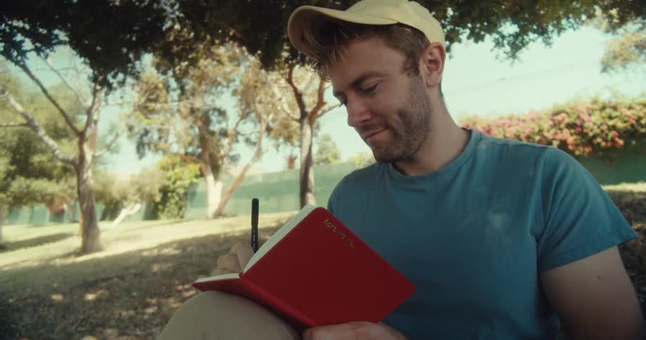 A man in a yellow cap and blue shirt sits in a park on a sunny day writing in a notebook under a tree