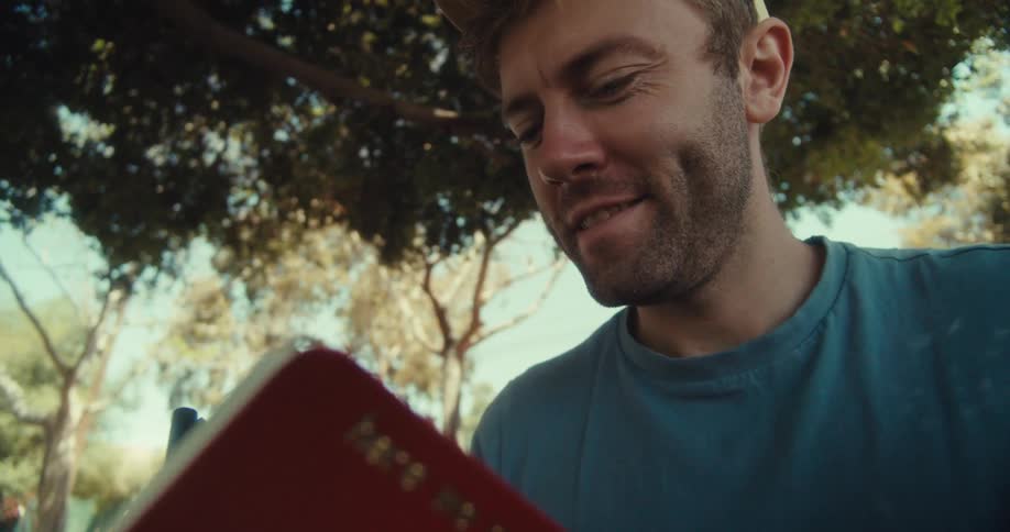 Man in cap with happy facial expression sitting under a tree and writing