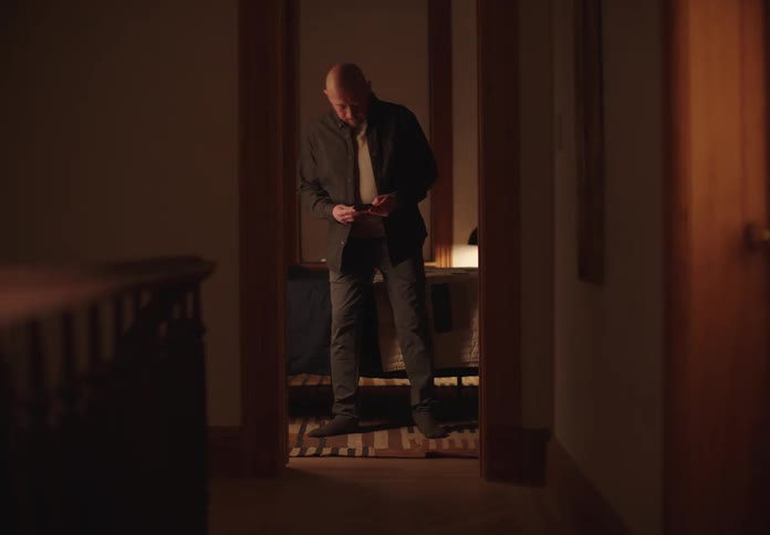 A man walks into his bedroom sits on the bed and scrolls through his phone while wearing a black shirt and jeans