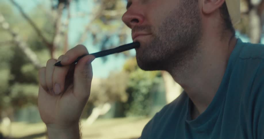 Man sitting outdoors putting pen in mouth and looking thoughtful