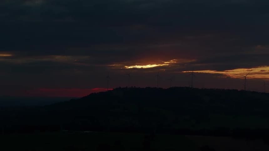 A dramatic sunset illuminates the cloudy sky over a dark hill lined with wind turbines