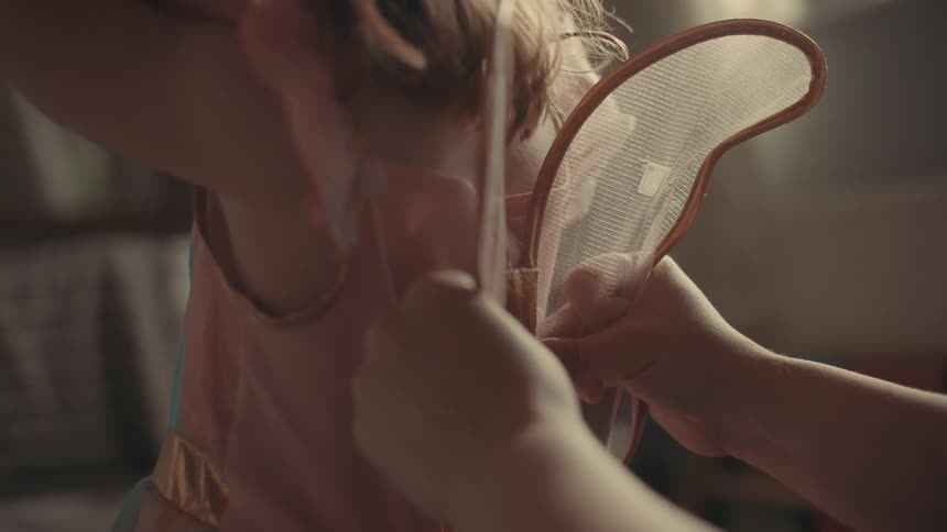 A mother clips a set of wings onto her daughters dress indoors