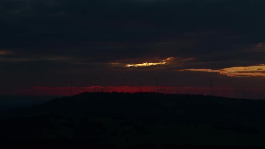 Wind turbines are silhouetted on a hilltop against a dark cloudy sky with a sliver of orange light on the horizon