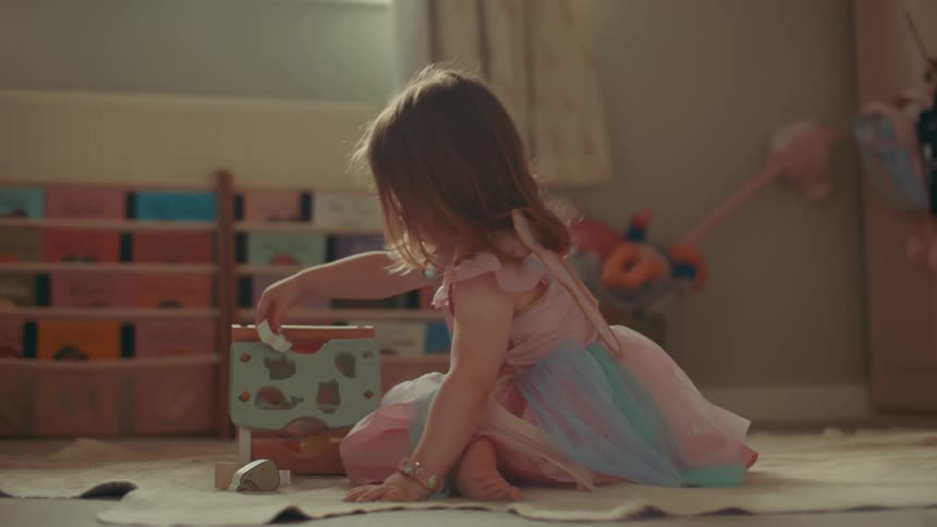 A young girl wearing a pink and blue dress with fairy wings plays with toys in a room