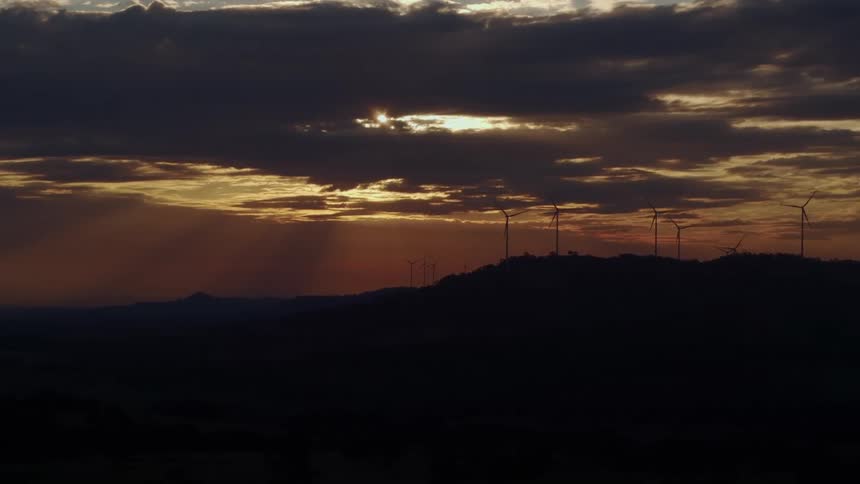 Windmills are on the mountain top back lit by a golden sunset through stormy clouds