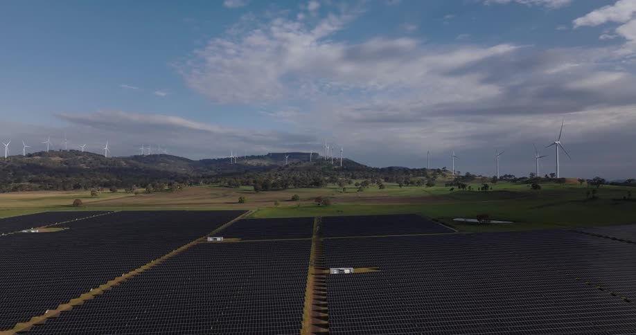 Wide aerial shot of a renewable energy farm with solar panels in front and wind turbines on distant hills under a partly cloudy sky