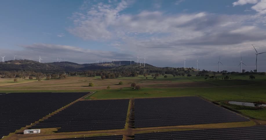 An aerial shot reveals large arrays of solar panels in the foreground with numerous wind turbines spinning on distant hills under a partly cloudy sky