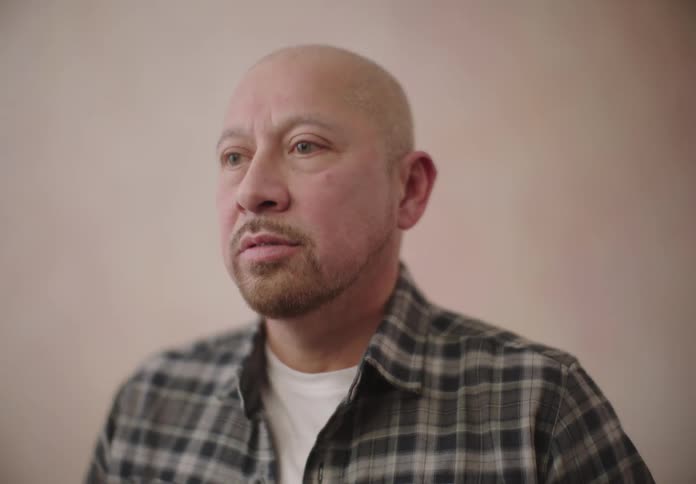 A bald man standing next to a wall adjusts the collar of his shirt