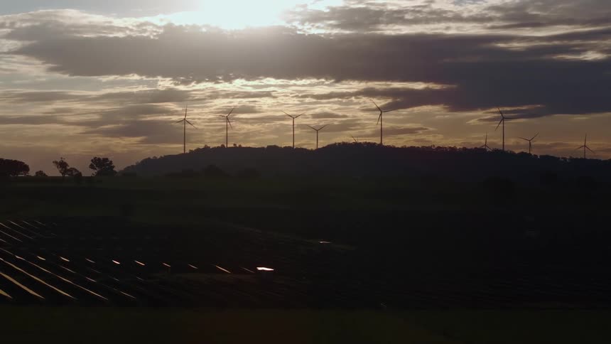 Wind turbines are silhouetted on a hill beneath a cloudy sky with rows of solar panels in the shadowed foreground as the sun shines through the clouds
