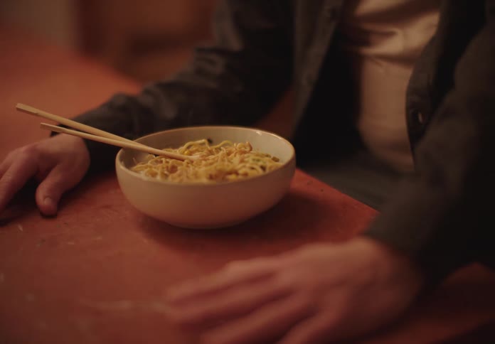 A man seats in front of a bowl of noodles with chopsticks resting on a table