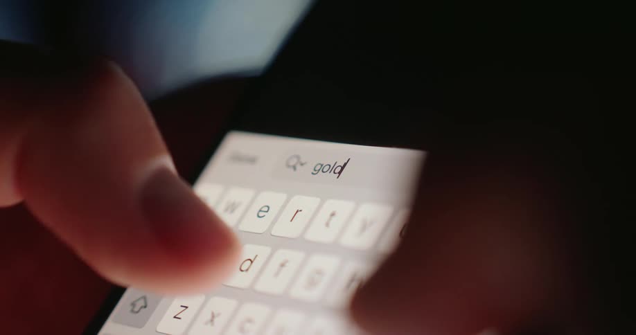 A closeup shot shows fingers typing the word gold into a search bar on a smartphone screen