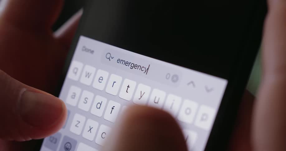Fingers type the word emergency on a brightly illuminated smartphone keyboard