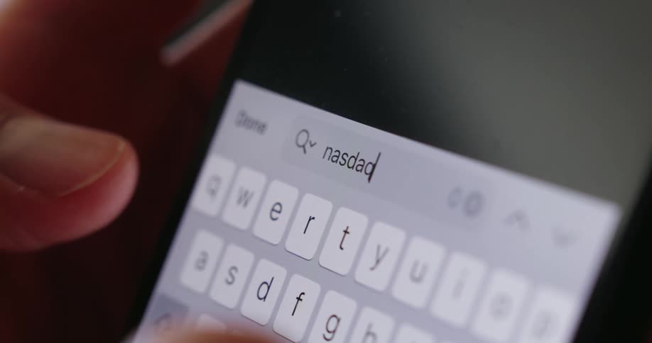 A persons finger holds a smartphone while typing nasdaq on it