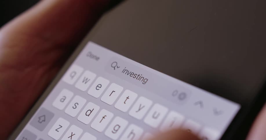 Fingers type the word investing on a brightly illuminated smartphone keyboard in a dimly lit area