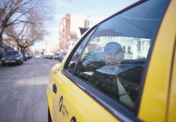 The face of a man who is inside a taxi looking out the window while it is moving along a street full of cars parked on the sides