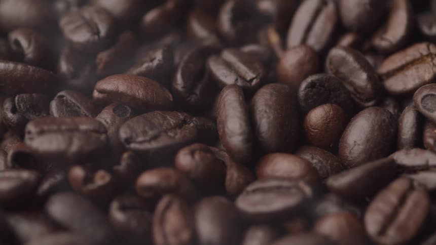 A closeup slowmotion shot captures dark roasted coffee beans with wisps of steam or smoke rising from them