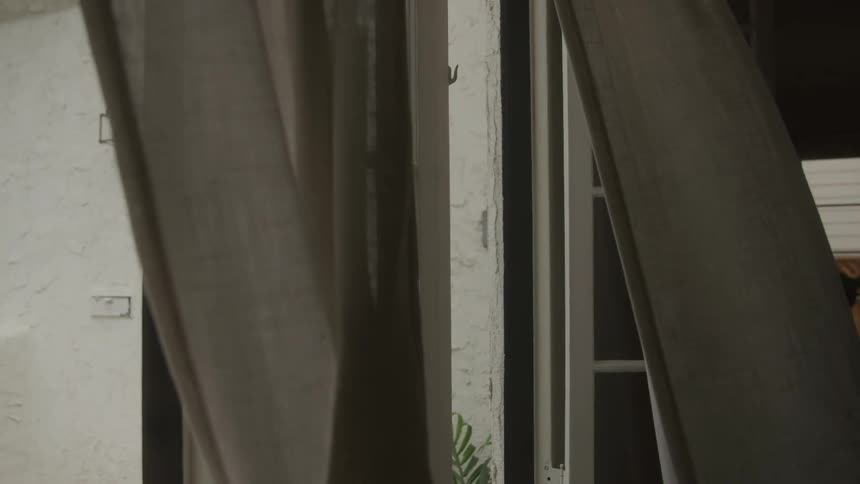 Sheer curtains billow gently in a dim room as wind blows through an open window