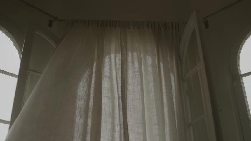 Wind blowing through an open arch shaped window disturbs the curtains during the daytime