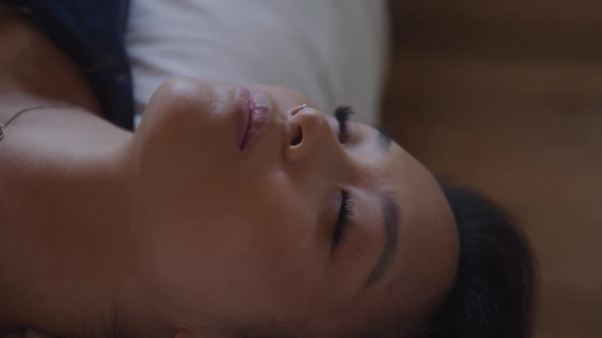 A closeup shot shows a womans face with her eyes closed lying on her back
