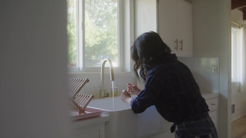 A distressed woman washes dishes at a kitchen sink then breaks down and rests her head in despair