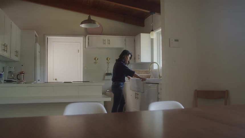 A woman stands alone in a quiet kitchen appearing sad and deep in thought while unloading the dishwasher