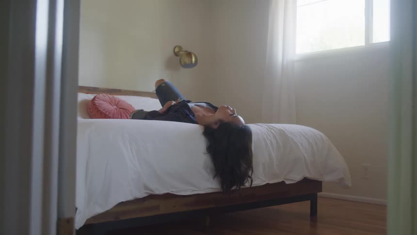 A woman lies on a bed in a softly lit bedroom looking out the window in a calm thoughtful mood