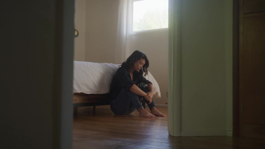 Woman sits on the floor near a bed during the daytime looking downcast