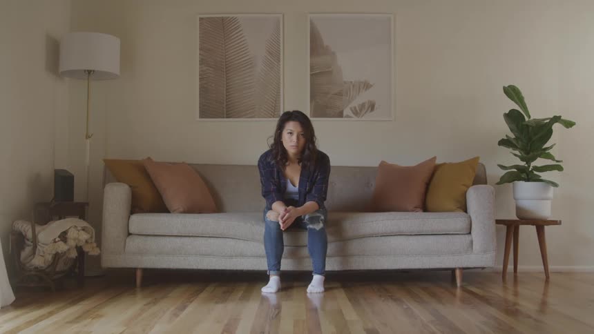 A young woman sits alone on a couch looking sad with a flower plant beside the couch at midday