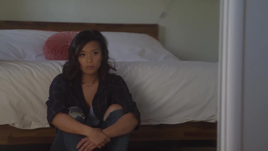 Woman sitting on the floor at the foot of the bed looks up with an upset face