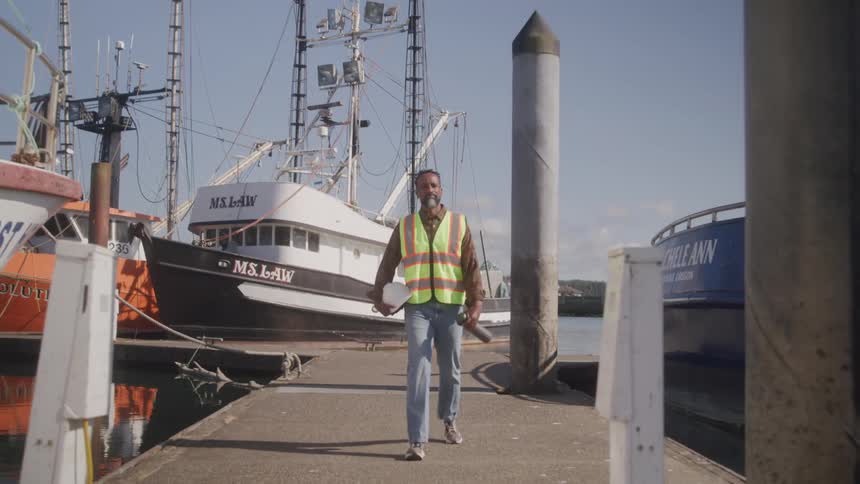 A black man wearing a green vest and holding a hard hat walks out of a boating dockyard filled with several boats at midday