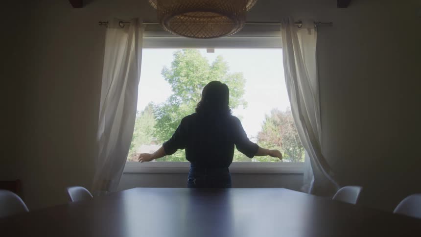 A silhouetted woman in a dining room opens the curtains to reveal a bright treefilled view outside the window